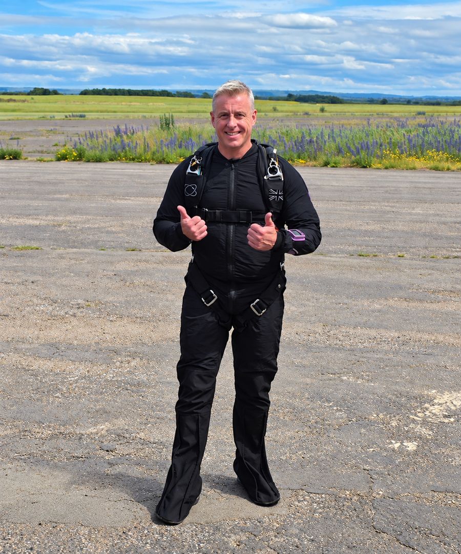 Nick on the drop zone in his skydiving kit, giving a double thumbs up